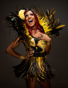 Joyful performer in a yellow-and-black feather costume, with a large yellow flower in her hair, smiling broadly against a dark backdrop.