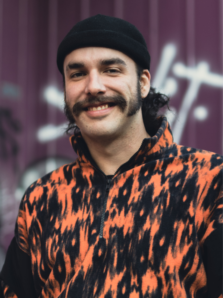 Portrait of a smiling man wearing a black beanie and an orange and black patterned fleece, standing in front of a purple graffiti wall.