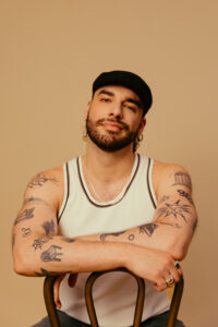 Bearded man in a white tank top and black cap resting his arms on gym rings, against a beige background, showing tattoos on his arms.