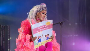 Drag queen in a bright pink outfit reads a colorful children's book on stage, microphone nearby.