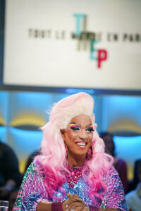 Drag queen with a long pink wig and sparkling, multicolored outfit smiling at a show.