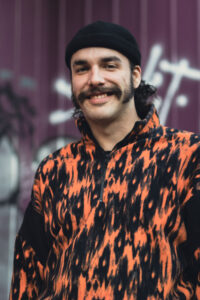 Smiling man wearing a black beanie and orange-black patterned jacket, standing in front of a purple graffiti wall.