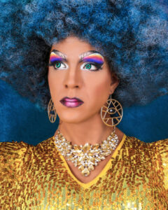 Portrait of a person with voluminous blue hair, bold rainbow eyeshadow, and teal contact lenses wearing gold jewelry and a sequined gold top.