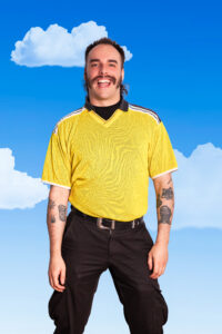 Smiling man in a yellow sports jersey outdoors with a blue sky and clouds behind him, tattoos visible on his arms.